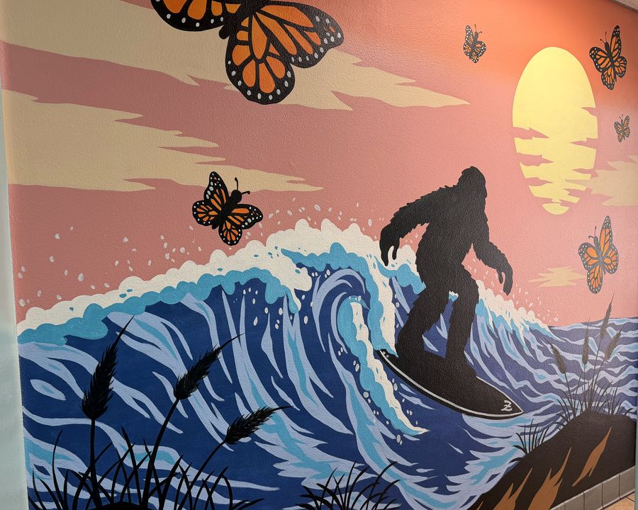 The mural on the wall inside: Bigfoot surfing a breaking wave under a sunset, monarch butterflies over the water.